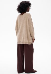 Beżowy oversize sweter o teksturowym wykończeniu, z opadającymi ramionami i bocznymi rozcięciami, w połączeniu z szerokimi brązowymi spodniami i butami na obcasie.