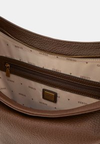 Interno della borsa in pelle marrone con texture a granuli, dotato di una tasca con zip e foderato con un motivo che ripete il logo "GUESS" e piccoli triangoli.
