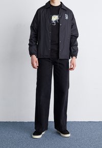 Mustat nailonitakki, jossa on nepparit ja joustavat hihansuut, yhdistetty mustaan graafiseen t-paitaan ja mustiin leveälahkeisiin housuihin. Valkopohjaiset lenkkarit.