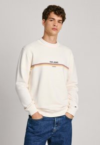 Sudadera crema de manga larga, con escote redondo y una franja horizontal multicolor. Presenta un logo impreso en el pecho. Combinada con jeans azules.
