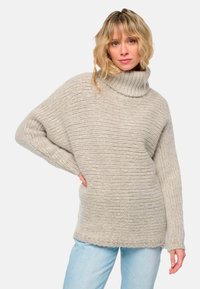 Frau mit blondem, welligem Haar, die einen dicken, gerippten beigen Rollkragenpullover und hellblaue Jeans trägt und mit einer Hand in die Hüfte posiert.