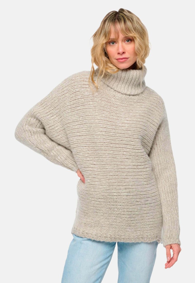 Frau mit blondem, welligem Haar, die einen dicken, gerippten beigen Rollkragenpullover und hellblaue Jeans trägt und mit einer Hand in die Hüfte posiert.