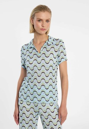 Frau mit kurzem blondem Haar, die ein passendes kurzärmeliges Oberteil und eine Hose mit blau-grün-schwarz gewelltem Muster trägt, steht vor einem weißen Hintergrund.