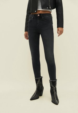 Vrouw draagt zwarte skinny jeans met rafelige zomen, zwarte halfhoge laarzen, een witte cropped top en een zwarte jas met rits.