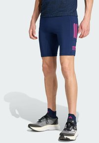 Homme portant un short de sport bleu marine avec des rayures roses et du texte, des chaussettes bleues navy et des chaussures de course grises sur un fond clair.