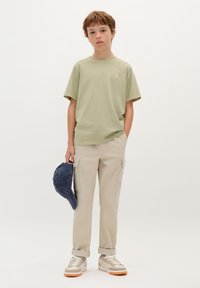 Lichtgroene katoenen T-shirt met een klein logo, gecombineerd met beige cargobroeken en grijze sneakers, terwijl hij een marineblauwe pet vasthoudt. Eenvoudige, casual outfit.