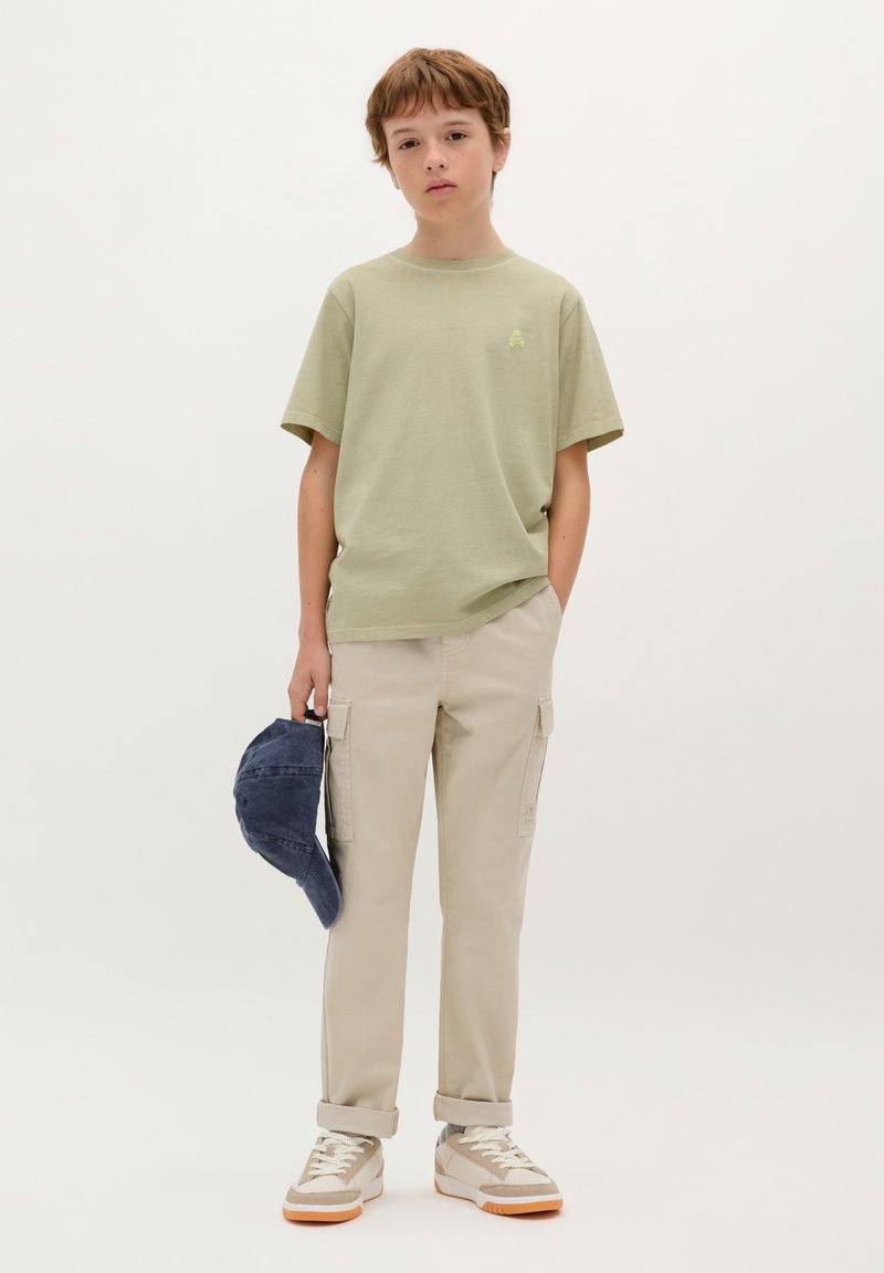 Lichtgroene katoenen T-shirt met een klein logo, gecombineerd met beige cargobroeken en grijze sneakers, terwijl hij een marineblauwe pet vasthoudt. Eenvoudige, casual outfit.