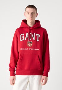 Sudadera roja de tela suave con bolsillo canguro, gran texto en blanco "GANT" y un pequeño parche con el logo. Cordones blancos en la capucha.