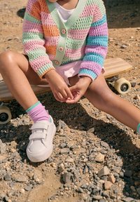 Un enfant portant un cardigan à rayures pastel et des chaussures blanches est assis sur un sol rocheux avec un skateboard à côté.
