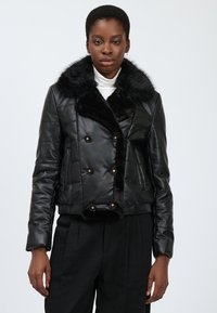 Veste en cuir noir courte avec un col en fausse fourrure, présentant un design à double boutonnage et des accents de boutons dorés. Texture lisse et détails matelassés.