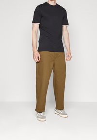 Zwart T-shirt met witte en rode gestreepte manchetten, gecombineerd met losse bruine broek en grijze sneakers met witte zolen en accenten.