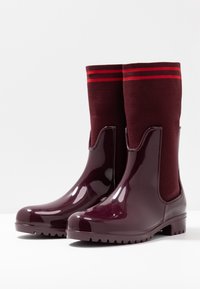 Un par de botas de lluvia de tobillo de un burdeos oscuro brillante con puños de punto que cuentan con dos rayas rojas en la parte superior, sobre una superficie blanca.