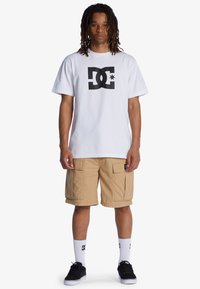 Bílý bavlněný tričko s černým logem DC, doplněné béžovými cargo šortkami. Černé tenisky a bílé ponožky s logem dotvářejí outfit.