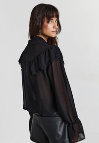 Frau mit langen dunklen Haaren, die eine durchsichtige schwarze Bluse mit Rüschendetails und schwarze Lederhosen trägt, und über die Schulter zu einem einfarbigen Hintergrund schaut.
