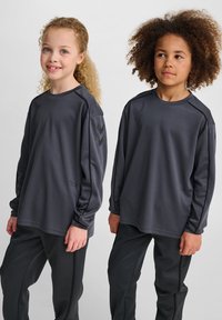 Två barn i grå sportiga långärmade tröjor och matchande byxor står sida vid sida och ler svagt mot en neutral bakgrund.