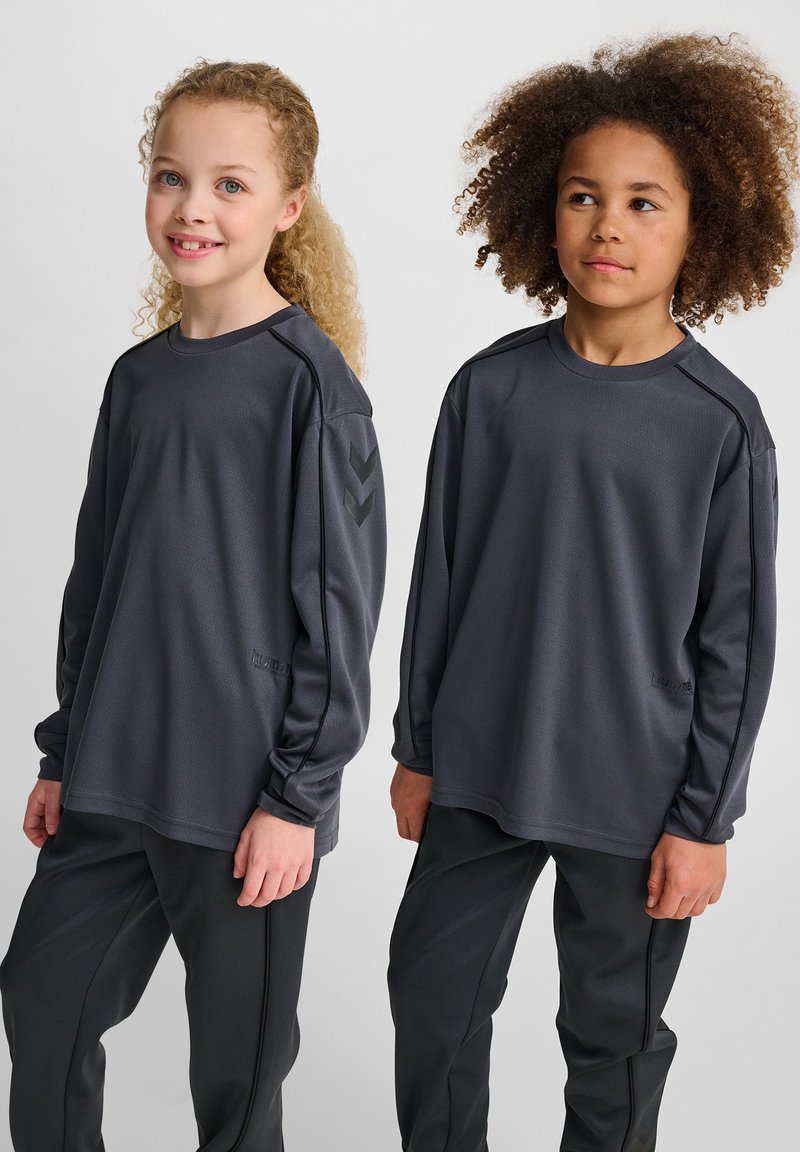 Två barn i grå sportiga långärmade tröjor och matchande byxor står sida vid sida och ler svagt mot en neutral bakgrund.