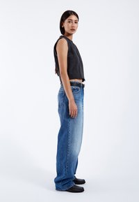 Zwarte gekorte mouwloze top, lichtblauwe baggy jeans met vervaagde accenten, en zwarte platte schoenen; model staat in profiel tegen een witte achtergrond.