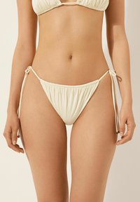 Krämfärgade bikiniunderdelar med veckad textur, sidoknutar och låg midja. Har mjukt tyg med minimal hårdvara.