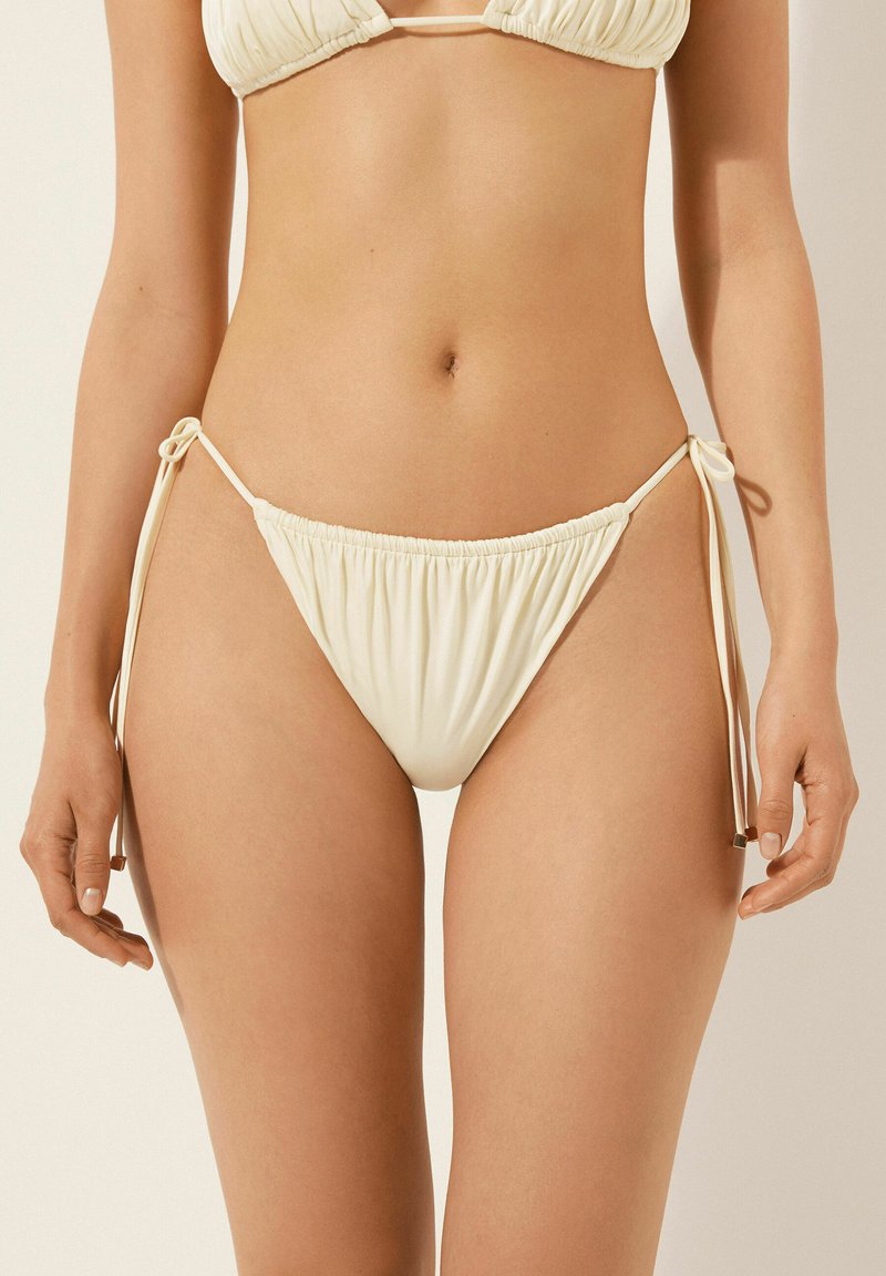 Kvinde iført cremefarvet bikiniunderdel med bindsler i siderne, vist fra midt på torsoen til øvre lår mod en neutral baggrund.
