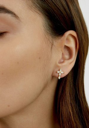 Close-up of a closed-eye person wearing a gold cross-shaped earring with clear gemstone accents on the left ear.