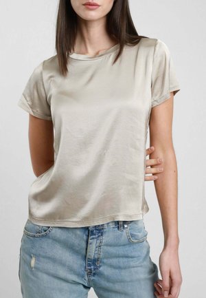 Donna che indossa una maglietta beige in satin a maniche corte infilata leggermente in un paio di jeans azzurri strappati, in piedi davanti a uno sfondo grigio semplice.