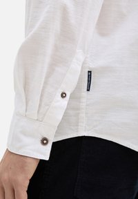 Chemise blanche à manches longues texturée avec des poignets boutonnés et une petite étiquette logo. Présentée avec un tissu sombre en dessous, mettant en avant le look superposé.