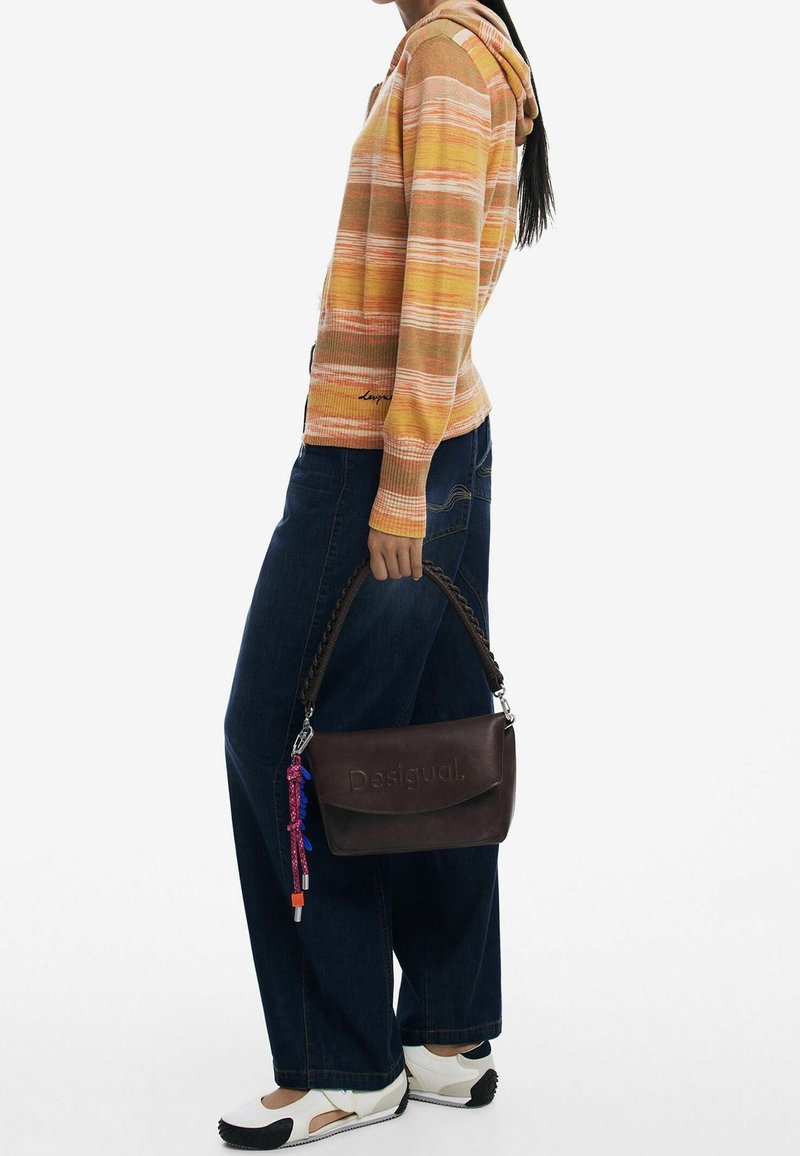 Personne portant un pull à capuche rayé jaune et orange, un jean large foncé, des baskets blanches, tenant un sac à main marron foncé avec une anse tressée.