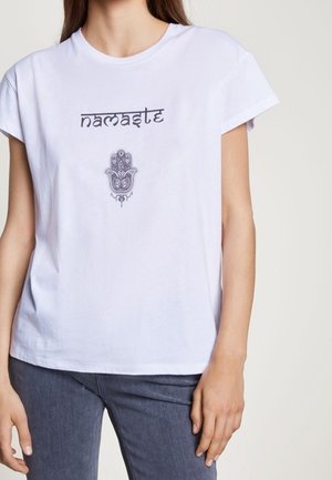 Kvinde iført hvid t-shirt med ordet "namaste" og et dekorativt håndsymbol trykt på fronten, kombineret med mørke jeans.