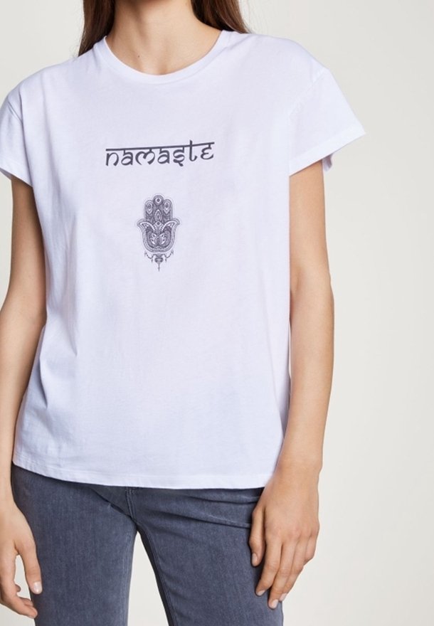 Femme portant un t-shirt blanc avec le mot "namaste" et un symbole décoratif de main imprimé à l'avant, associé à un jean foncé.