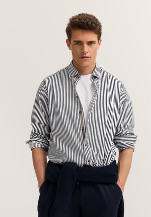 Junger Mann, der ein weiß- und schwarzgestreiftes Hemd über einem weißen T-Shirt trägt, mit einem marineblauen Pullover um die Taille gebunden.