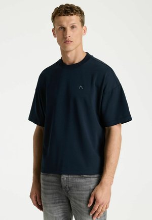 Young man with curly hair wearing a plain dark navy oversized t-shirt and light faded gray jeans, standing against a white background.