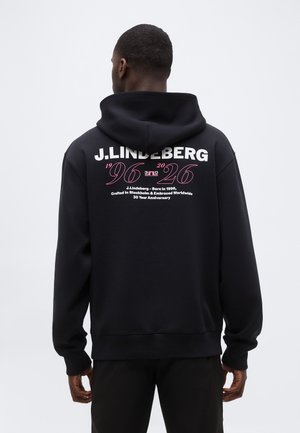 Mann, von hinten gesehen, trägt schwarzen J.Lindeberg-Hoodie mit "96 26" und Jubiläumstext in Weiß und Pink auf dem Rücken gedruckt.