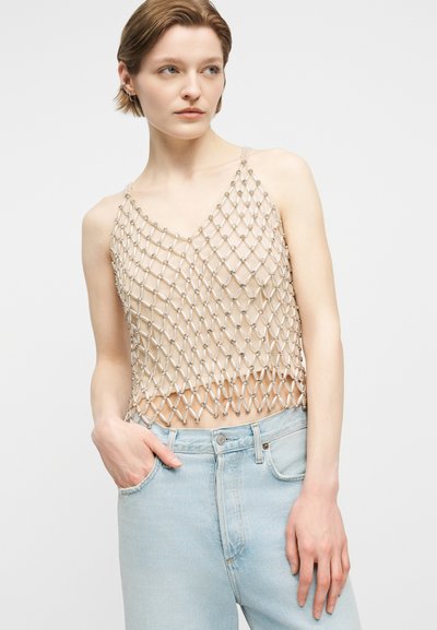 Νεαρή γυναίκα που φοράει μπλούζα από μπεζ δίχτυ με λεπτομέρειες από χάντρες πάνω σε γυμνό υπόστρωμα, συνδυασμένη με ανοιχτό μπλε τζιν, στέκεται μπροστά σε λευκό φόντο.