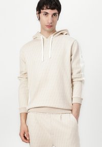 Felpa beige con un motivo geometrico testurizzato, cappuccio con coulisse e polsini a coste. Abbinata a pantaloni jogger leggeri coordinati.