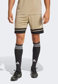 Béžové sportovní šortky s černými a zlatými pruhy, v kombinaci s vysokými černými ponožkami s bílými pruhy a logem Adidas.