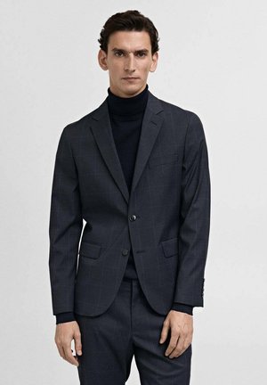 Mörkgrå skräddarsydd blazer med ett subtilt rutmönster, enkel knappstängning, slagna kragar och två framfickor. Bärs över en svart polo.