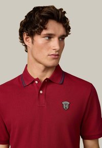 Czerwona koszulka polo z fakturowanej tkaniny, z kołnierzykiem z niebieskimi akcentami oraz naszywką z logo na lewej piersi.