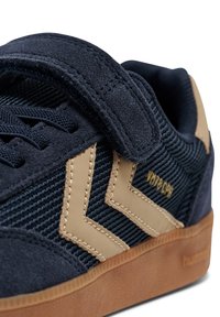 Granatowy sneaker z teksturowanym zamszem i siateczką, z beżowym skórzanym wzorem w zygzak. Wyposażony w pasek na rzep i podeszwę z gumy naturalnej, tworzy kompletny wygląd.