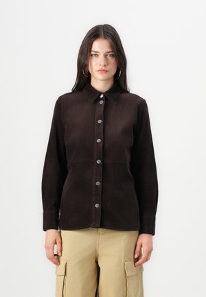 Chemise en velours côtelé marron avec un col, fermeture à boutons à l'avant et manches longues. Présente une texture à côtes verticales et des boutons en métal contrastants.