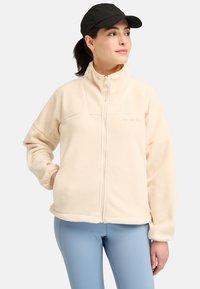 Light beige fleece jacket with a high collar, full zipper, and pockets. Features soft texture and subtle branding on the chest.
