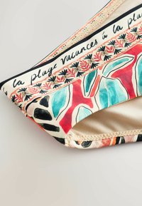 Közelkép színes bikini alsóról, virágos és absztrakt mintákkal, valamint francia szöveggel a szövet szélén: „la plage vacances á la plage”.