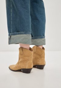Cizme scurte din daim bej, cu un design cu vârf curbat și cusături înclinate. Toc mic de bloc și talpă de culoare maro închis vizibile deasupra blugilor denim rulați.