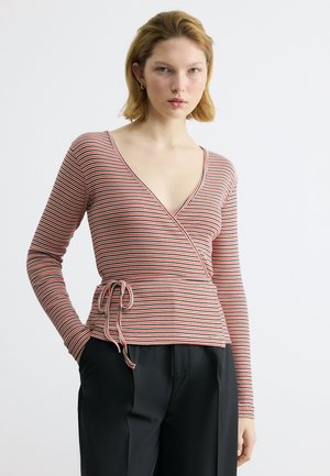 Γυναίκα με κοντά ξανθά μαλλιά, φορώντας ένα κόκκινο, λευκό και μαύρο ριγωτό πλεκτό πουκάμισο δεμένο στη μέση και μαύρο παντελόνι, στέκεται μπροστά σε λευκό φόντο.