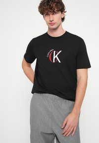 Czarny bawełniany T-shirt z białym i czerwonym logo Calvin Klein. Okrągły dekolt, krótkie rękawy i luźny krój, w zestawieniu z szarymi spodniami.
