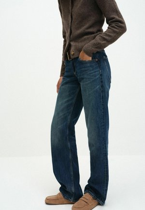 Person iført mørkeblå jeans, brun cardigan og lysebrune ruskindslofers, med den ene hånd i lommen mod en ensfarvet hvid baggrund.