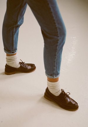 Scarpe in pelle marrone con lacci, suola in gomma, indossate con calzini color crema, abbinate a jeans in denim blu arrotolati.