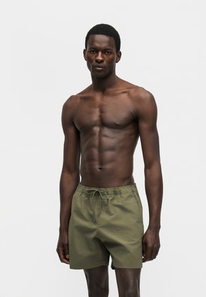 Γυμνασμένος άνδρας με σκούρο δέρμα στέκεται σε χαλαρή στάση, φοράει μαγιό χακί χρώματος, με απλό φωτεινό φόντο.