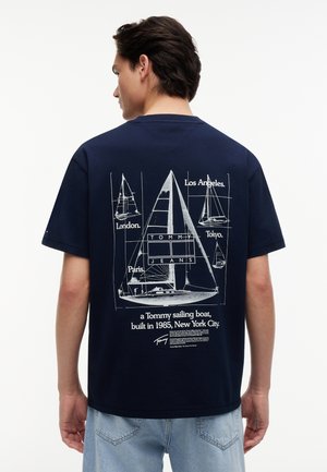 Άνδρας με μπλε μπλουζάκι ναυτικού χρώματος που φέρει σχέδιο λευκού ιστιοπλοϊκού σκάφους και ονόματα πόλεων: Λος Άντζελες, Λονδίνο, Παρίσι, Τόκιο, και κείμενο σχετικά με το σκάφος Tommy του 1985.