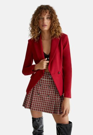Blazer rosso realizzato in tessuto con finitura satinata, abbinato a una mini gonna a scacchi rossi e neri. Presenta un design doppiopetto e dettagli in bottoni.