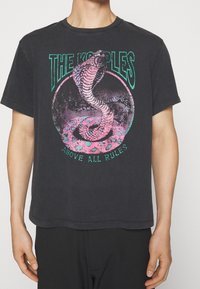 Camiseta de algodón negra con un gráfico de una serpiente en rosa y verde azulado dentro de un diseño circular. El texto dice "LOS KROUPLES POR ENCIMA DE TODAS LAS REGLAS."